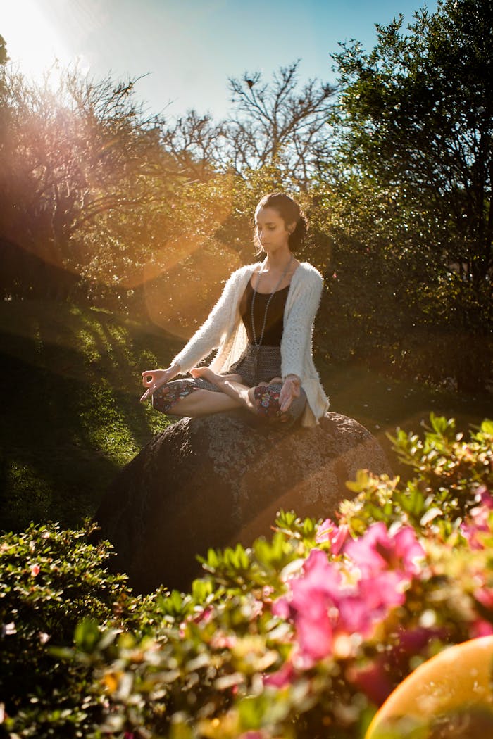 Woman in tranquil meditation pose outdoors, bathed in sunlight, surrounded by lush greenery.